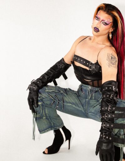 Saltina Shaker wearing a black sheer underbust corset, black strappy crop top with feather armbands, and grey denim jeans. She's posing in a squat position with her head tilted to the right.