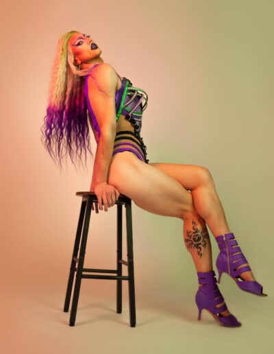 Saltina Shaker wearing a green, purple, and black strappy leotard with matching purple strappy heels. She's posing on a stool with her legs crossed and head tilted back.