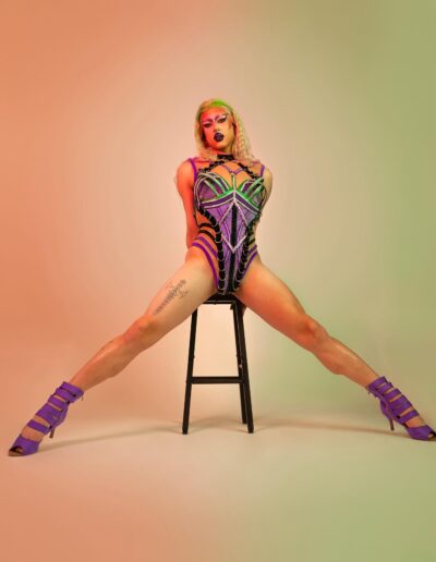 Saltina Shaker wearing a green, purple, and black strappy leotard with matching purple strappy heels. She's posing with her legs spread to the side, supporting herself with her hands on a stool behind her.