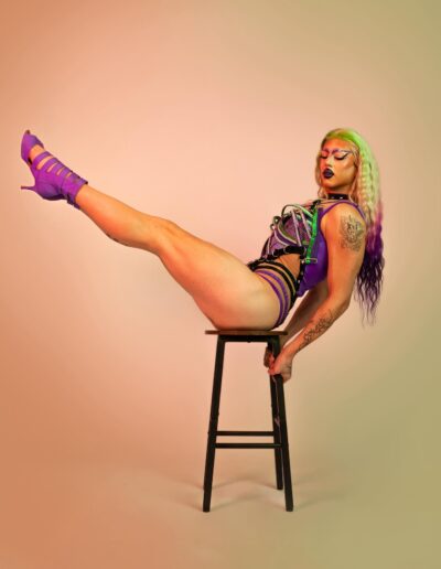 Saltina Shaker wearing a green, purple, and black strappy leotard with matching purple strappy heels. She's posing on a stool with her body in a V shape.