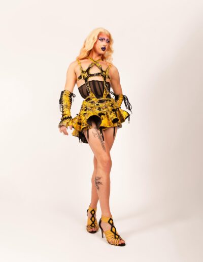 Saltina Shaker wearing a black and yellow strappy outfit over a black sheer underbust corset. She's posing with her right leg in front of her left and left hand behind her back.