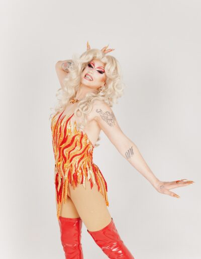 Sunshine Glitterchild in a red, yellow, and orange flame outfit, posing dramatically with her right hand on her forehead and left hand behind her.