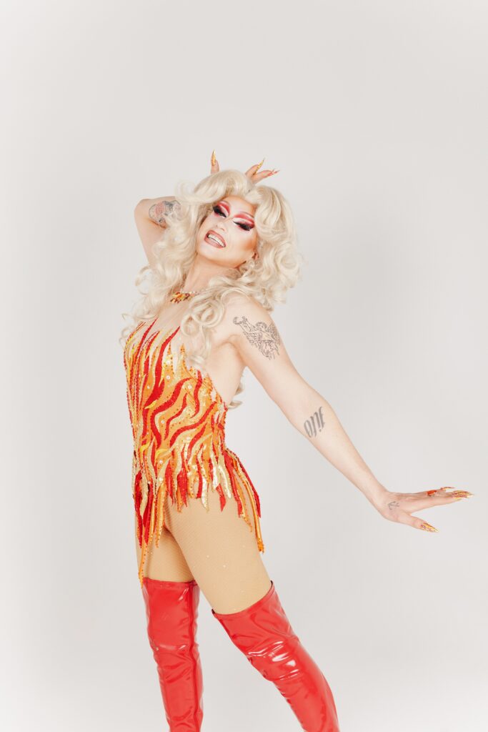 Sunshine Glitterchild in a red, yellow, and orange flame outfit, posing dramatically with her right hand on her forehead and left hand behind her.