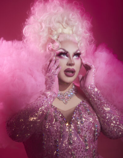 Beth wearing a pink dress with iridescent rhinestones on the bodice and sleeves with ruffled shoulders, touching her face and gazing into the distance.