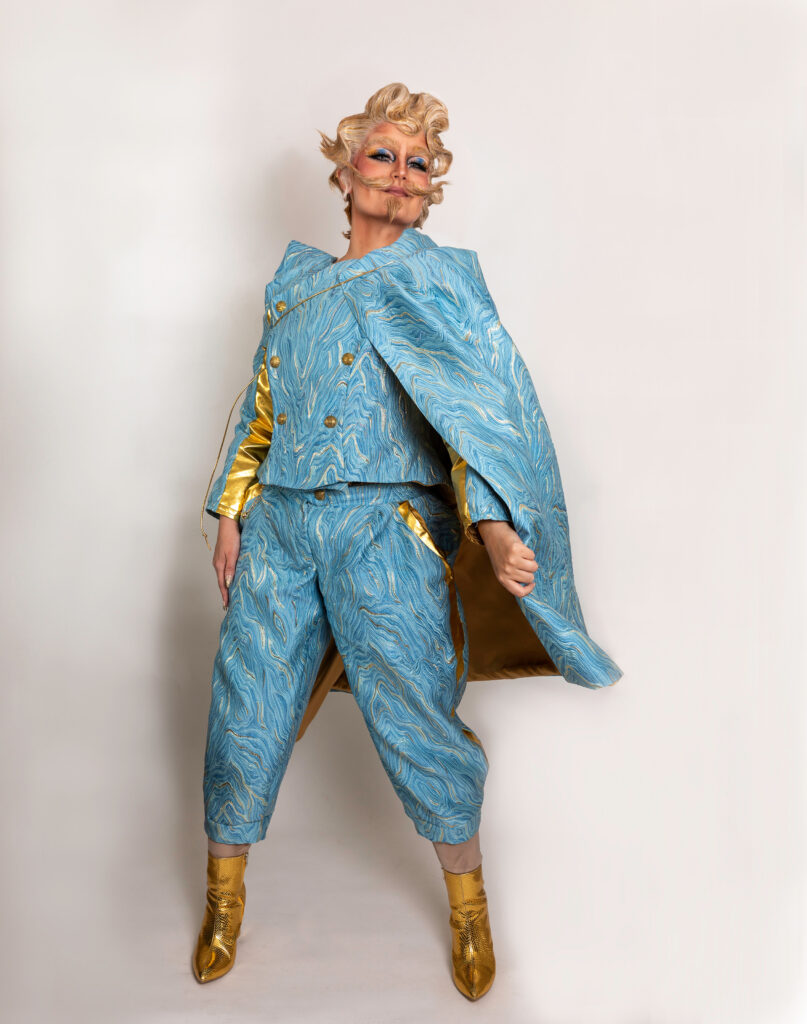 Cyril Cinder posing dramatically in a blue marbled suit with matching cape and gold accents.