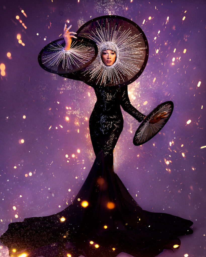 Bombae-Firework Beth posing demurely in a glittery champagne pink fitted gown, her hands up by her shoulders and face.