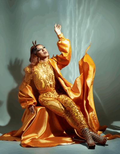 Mya Foxx in a lavish gold bodysuit and robe, flinging the robe while sitting.