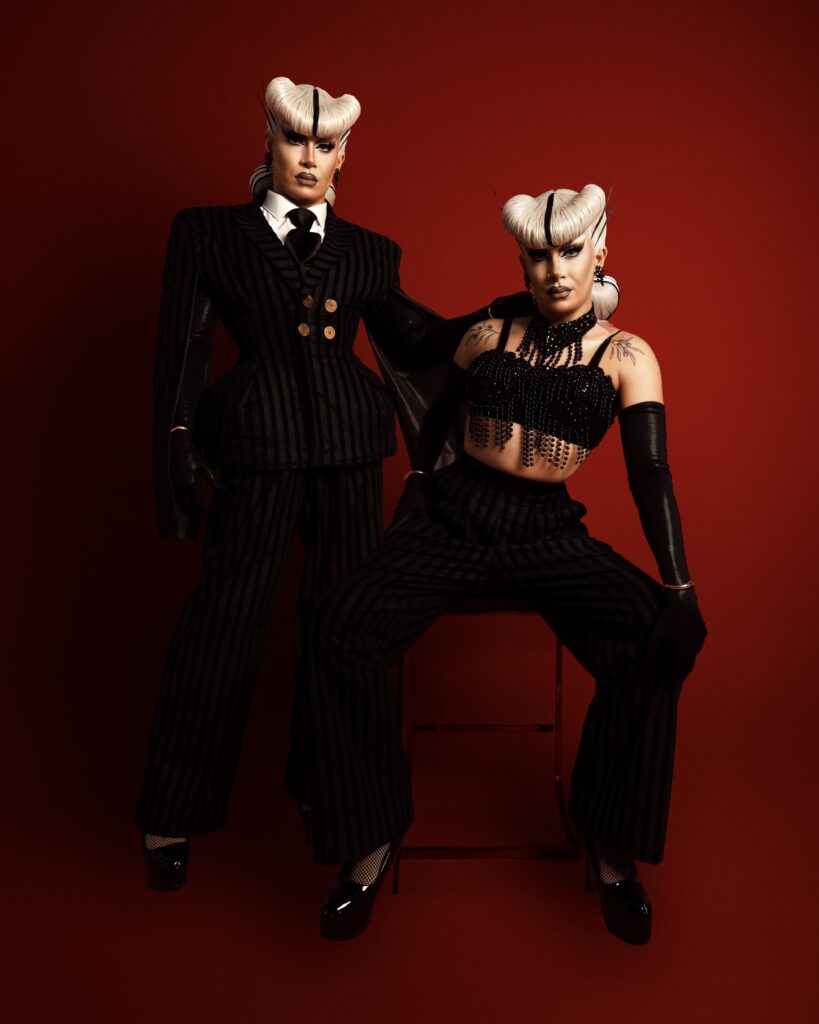 Photo of two Vanity Affairs, one standing in a suit and jacket, one sitting and wearing a cropped tank top and black gloves with trousers.