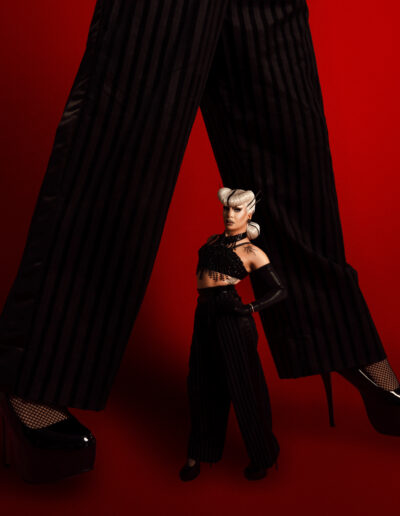 Vanity Affair in a black cropped tank top with gloves and suit trousers, posing in front of a background of her legs.