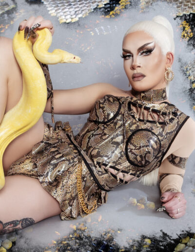 Vanity Affair in a snakeskin bodysuit posing on the floor with a leucistic ball python.