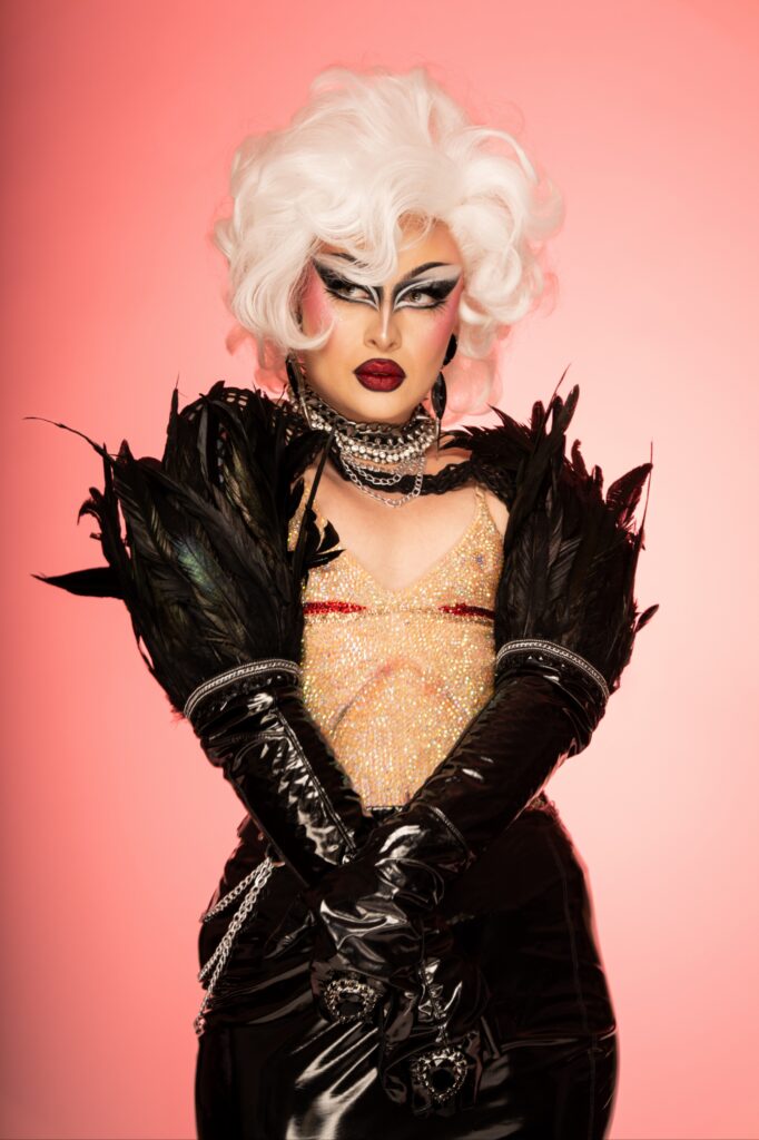 Phoenix Black wearing a white and gold silk gown, seated on a stool with her left hand on her hip and right hand next to her face.