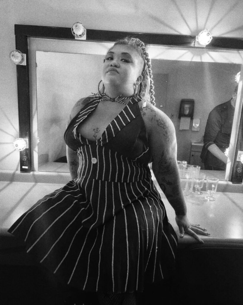 Phoenix Black wearing a white and gold silk gown, seated on a stool with her left hand on her hip and right hand next to her face.